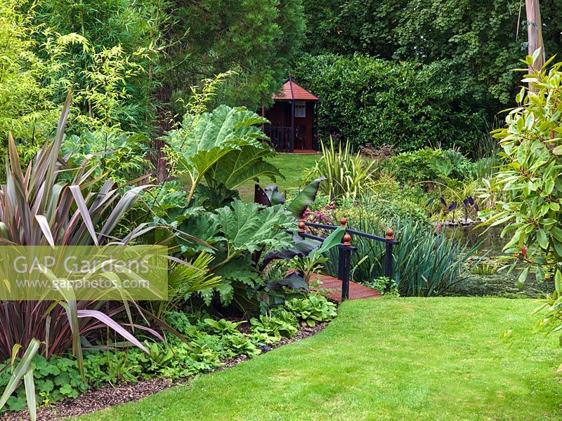 Bridge over pond. Borders with Phormium 'Rainbow Queen', Gunnera manicata, Phyllostachys nigra and canna. 