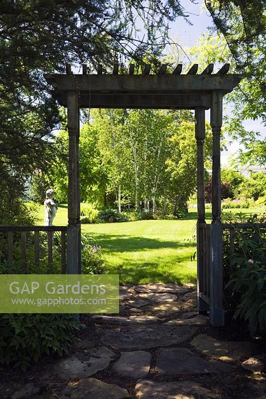 Flagstone path through silhouetted wooden arbour leading to a sun drenched green grass lawn with Betula - Birch trees in backyard country garden in spring, Quebec, Canada