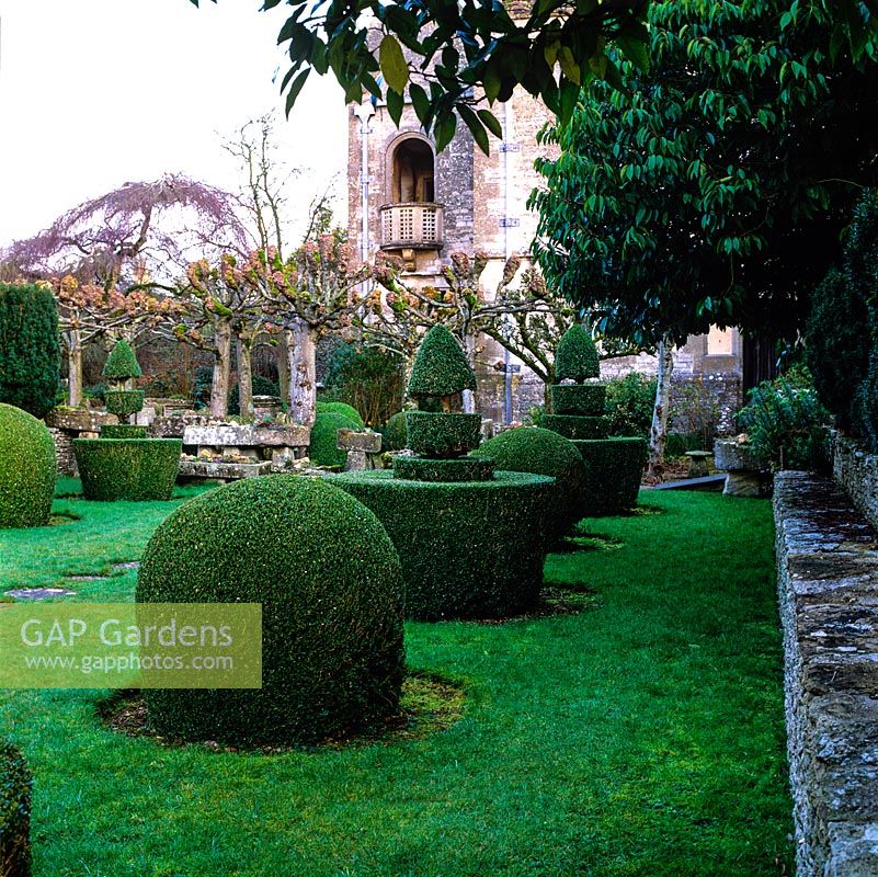 Topiary shapes in clipped box march across lawn leading to Arts and Crafts house flanked by stone troughs and avenue of limes.