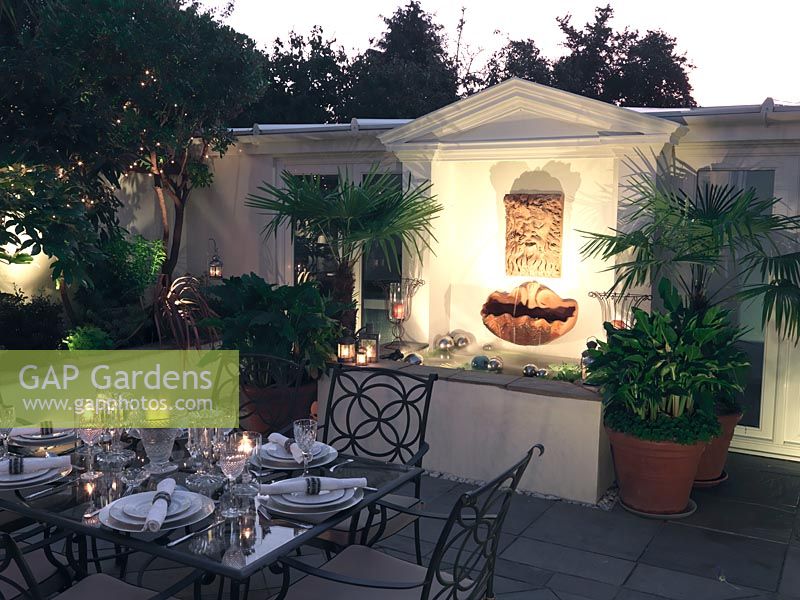 Walled outdoor room at night with illuminated wall fountain mask trickling water into raised pool flanked by pots of palms and arum lilies. Table laid for dinner.