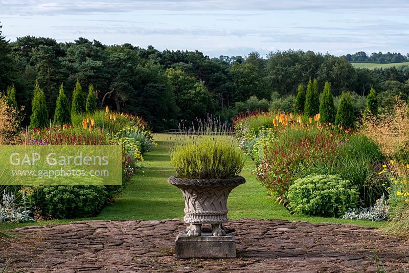 A decorative stone planter with lavender at the head of a grass path leading between twin herbaceous borders with repeated miscanthus, stipa and Thuja occidentalis 'Smaragd' - conifers. Set against backdrop of Utkinton Hills.