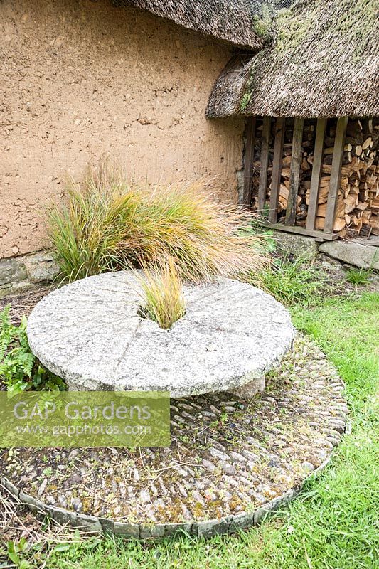 Millstone with grass growing up though it, with cob wall and thatched well head behind. Caervallack Farm, nr Helston, Cornwall, UK