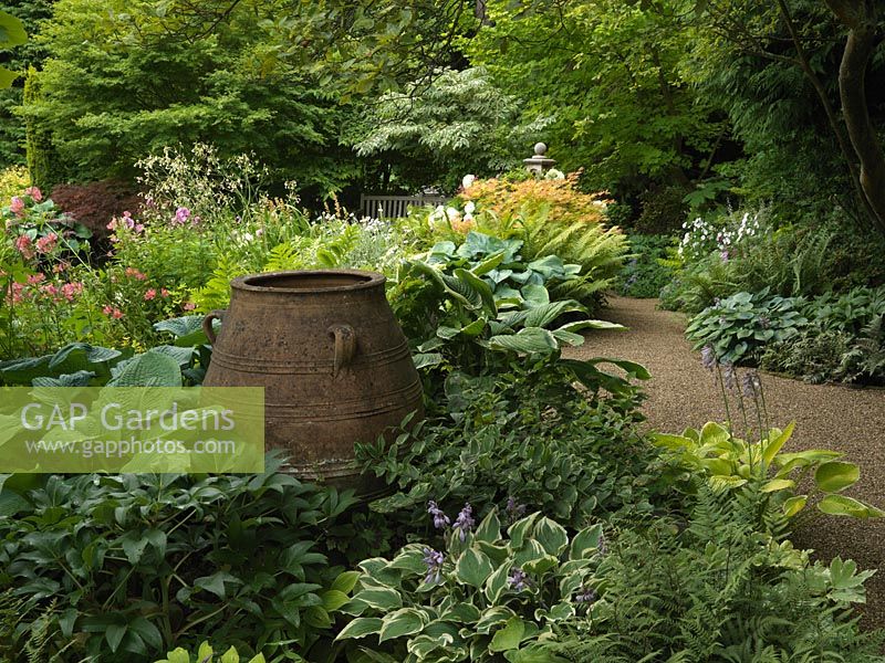 Cretan pot in circular leafy bed of Hosta 'Frosted Jade', hardy geranium, Helleborus argutifolius, Polygonatum odoratum 'Gilt Edge', Athyrium 'Ghost', saxifrage, Sanguinaria canadensis.