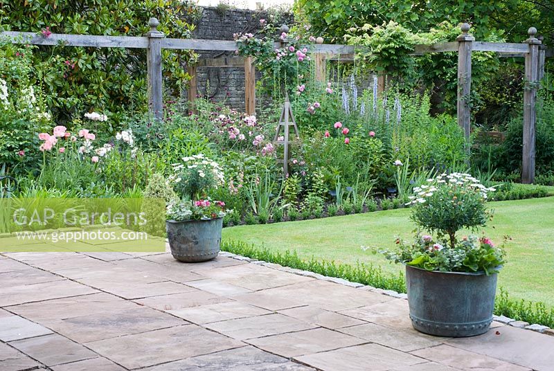 View of mixed Summer border under pergola, patio containers filled with Marguerites and bedding plants - Priory House, Wiltshire