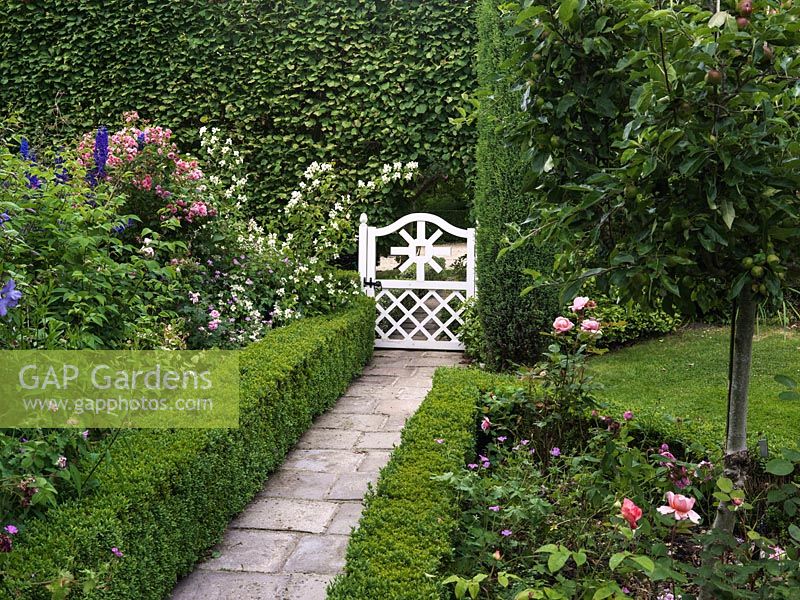 White gate opens onto stone path between box-edged beds of apples, clematis, delphinium, hardy geranium, philadelphus and roses, offset against tall beech hedge.