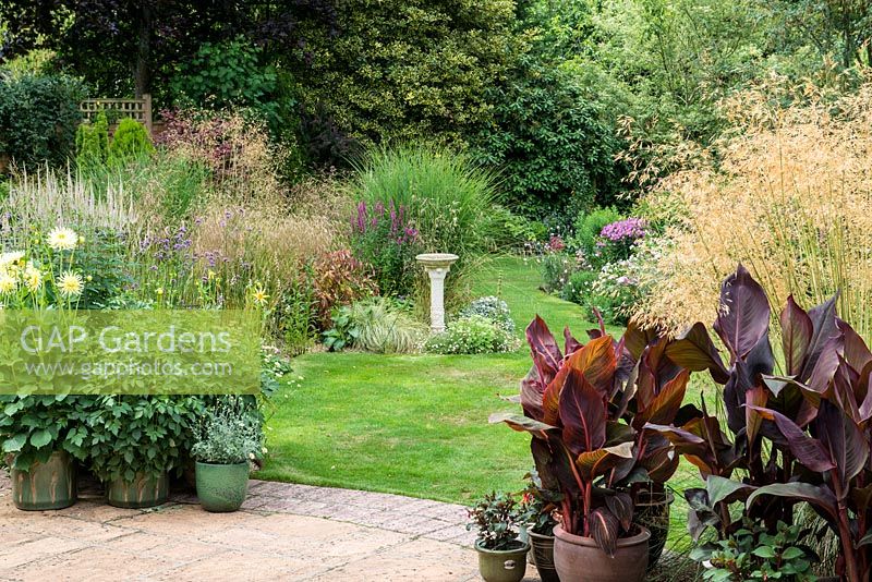 A bird bath at the front of a large herbaceous border providing a focal point from the patio. The patio containers are planted with Dahlia, Canna and Fuchsia.