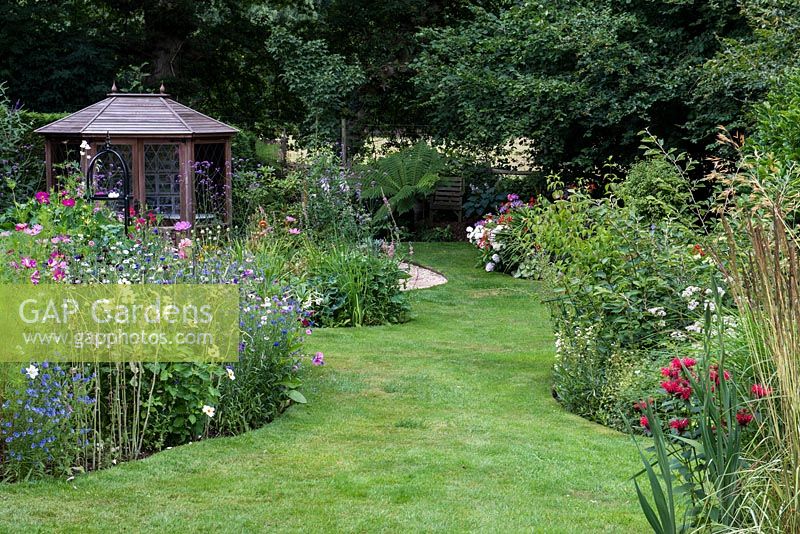 A grass path between a colourful wildflower border and a mixed herbaceous border, with a wooden summerhouse behind.