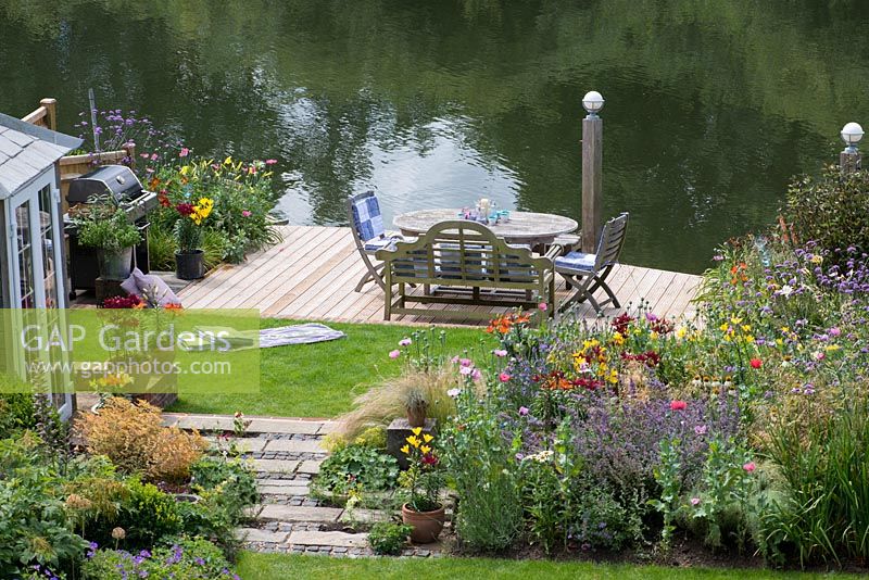 Bird's eye view of summerhouse by bed of catmint, coneflower, annual poppies and oriental lilies. Lawn leads to riverside deck edged in pots and beds of oriental lilies, daylilies, Verbena bonariensis, annual poppies, agapanthus and kniphofia.