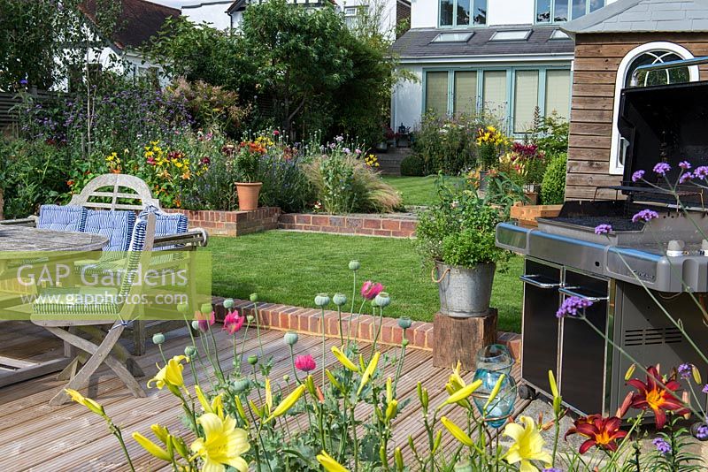 Riverside deck with barbecue, dining table and chairs, is edged in pots and bed of oriental lilies, daylilies, Verbena bonariensis and annual poppies. A step up leads to a lawn and raised bed.