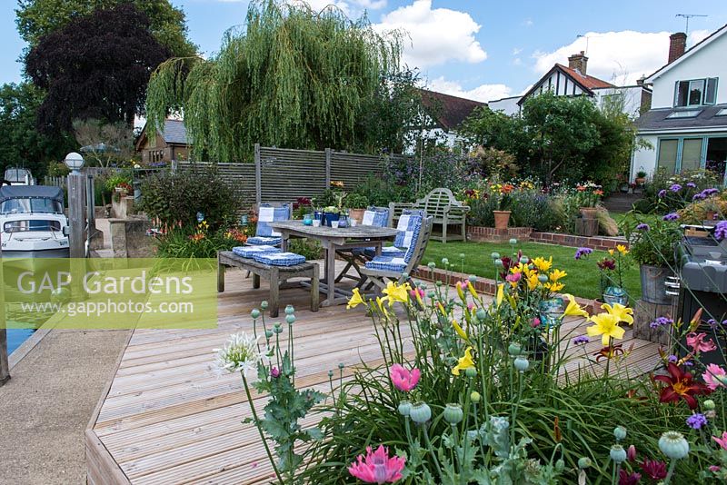 Riverside deck with dining table and chairs edged in pots and beds of oriental lilies, daylilies, Verbena bonariensis, annual poppies, agapanthus, dogwood and kniphofia.