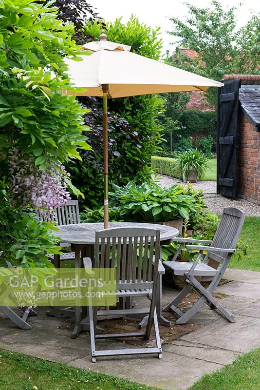 Wooden garden furniture on a small stone patio in front of a small container garden planted with Hosta 'Great Expectations', Hosta 'June' and at back Hosta 'Silk Road'.