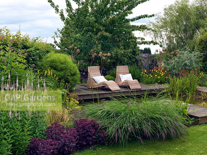 Wicker loungers on the pond side deck, behind a border including Veronicastrum virginicum 'Fascination',  Sedum telephium 'Purple Emperor' and Pennisetum alopecuroides,