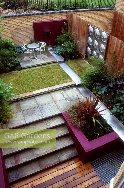 Modern city garden with a water spout into a stainless steel rill, surrounded by paving, patio, grass and stone steps. Designed by Paul Dracott.