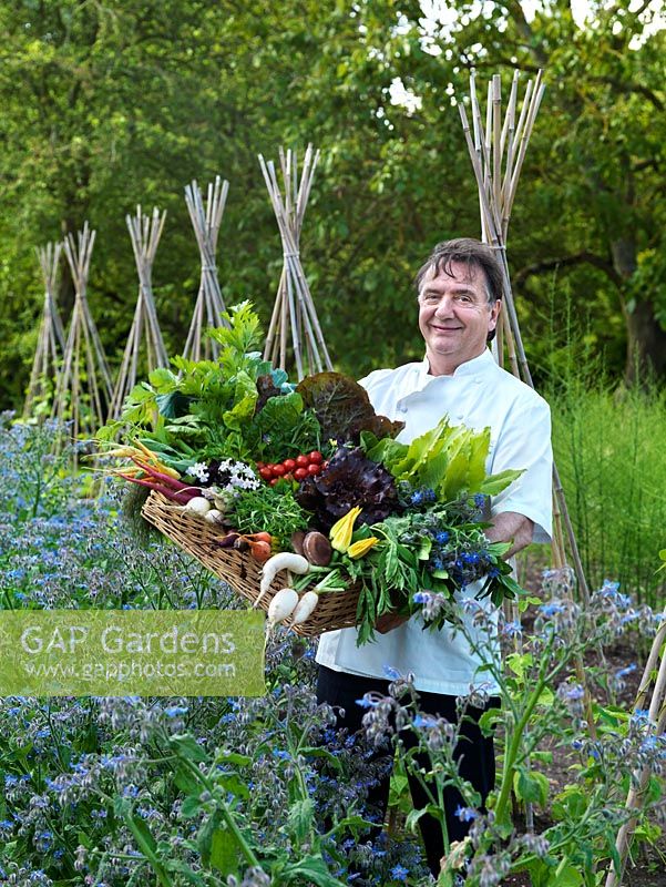Raymond Blanc stands in his organic vegetable garden against a backdrop of borage and can wigwams for beans, in his arms a box of fresh produce.