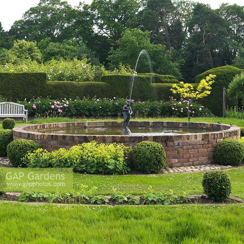 Formal raised circular pool with fountain, edged in box balls and Alchemilla mollis. Behind, peony cutting bed against old yew hedge.