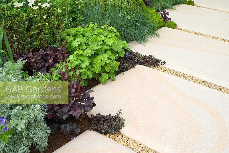 The Time In Between by Husqvarna and Gardena. Stone paving and gravel path with planting including: Artemisia absinthium, Heuchera obsidian, Ajuga reptans, Alchemilla mollis and Festuca glauca