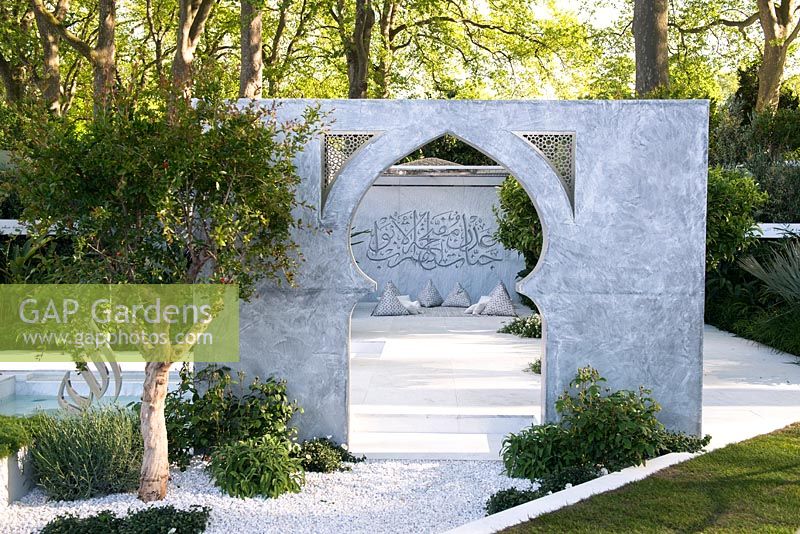 view of a seating area with cushions against marble wall with Arabic writing through an arch surrounded by Punica granatum, Cistus salviifolius, Alternanthera green. The Beauty of Islam - Chelsea Flower Show 2015