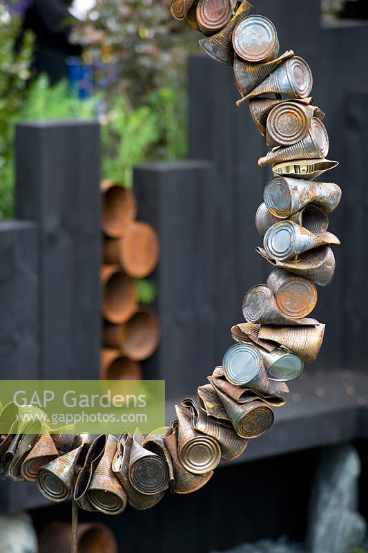 The Great Chelsea Garden Challenge. Sculpture made from crushed tin cans viewed against black wooden sleeper seat.