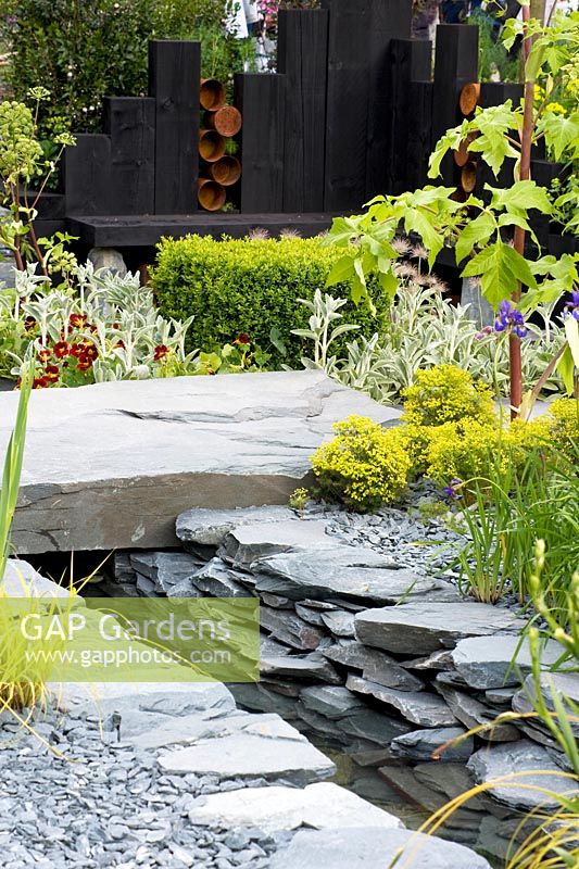 View across reclaimed stone hard landscaping with water channel toward black wooden seating area. Planting includes Angelica 'Ebony', Stachys, Euphorbia, Buxus and Nasturtium. The Great Chelsea Garden Challenge Garden, RHS Chelsea Flower Show, 2015