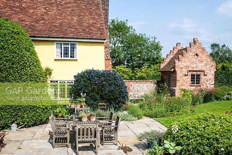 Terrace with wooden table and chairs, terracotta pots.  Laurus nobilis - bay tree, Ceanothus and perennials by a newly built brick garden house with crow stepped gables. Heveningham, June