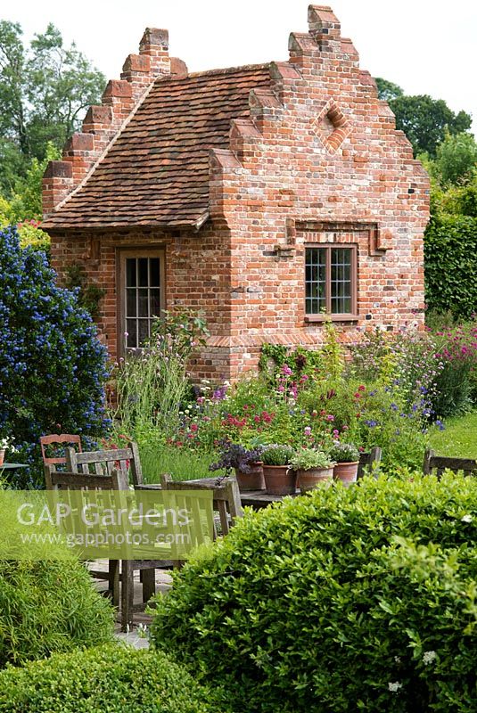 Terrace with wooden table and chairs, terracotta pots. Ceanothus and perennials by a newly built brick garden house with crow stepped gables. Heveningham, June