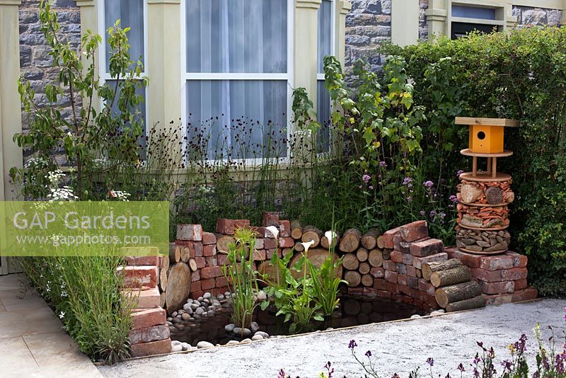 Front garden with pond made from recycled logs and bricks, and a recycled animal hotel in Community Street at Hampton Court Palace Flower Show 2015