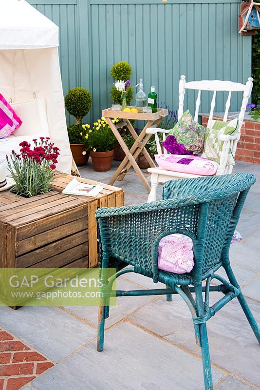 Just Retirement: A Garden For Every Retiree, view of seating area with blue painted wicker armchair and a table made from wooden crates against blue wall surrounded by clipped Buxus sempervirens in pots. Designer: Tracy Foster Sponsor:  Just Retirement Ltd