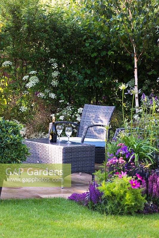 A garden by association, view of seating area with synthetic wicker armchairs and a table surrounded by Betula Jacquemontii, Anthriscus sylvestris, flowerbed with Cosmos,  Veronica longifolia 'Charlotte', Nepeta x faassenii 'Blue Wonder', Sambucus nigra porphyrophylla 'Eva' and pot with Pittosporum Tobira. Designer: Tina Vallis, MSGD