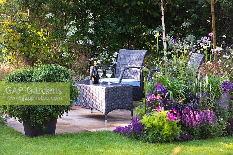 A garden by association, view of seating area with synthetic wicker armchairs and a table surrounded by Betula Jacquemontii, Anthriscus sylvestris, flowerbed with Cosmos, Veronica longifolia 'Charlotte', Nepeta x faassenii 'Blue Wonder', Sambucus nigra porphyrophylla 'Eva' and pot with Pittosporum Tobira - Designer: Tina Vallis, MSGD
