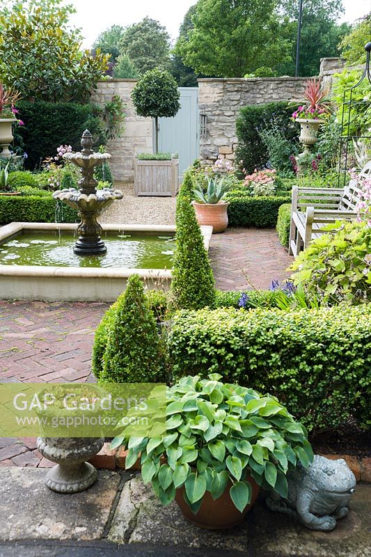 Formal courtyard garden behind Georgian terraced house with central water feature, box edged beds infilled with Japanese anemones, echinops and roses, box cones, stone urns, a standard bay and containers planted with succulents and hostas.