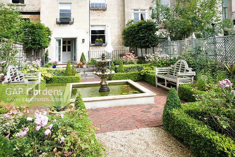 Formal courtyard garden behind Georgian terraced house with central water feature, box edged beds infilled with Japanese anemones, penstemons and roses, box cones and containers planted with succulents and agapanthus. Grey wooden trellis screens the garden on one side, pleached hornbeams screen a dining terrace directly adjoining the house.