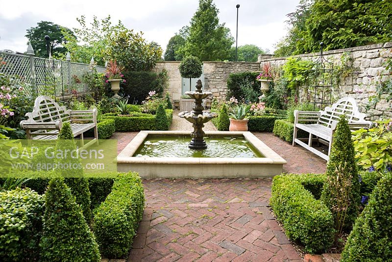 Formal courtyard garden with central water feature, Lutyens benches, box edged beds infilled with Japanese anemones, echinops and roses, box cones, stone urns with bright cordylines, a standard bay and containers planted with succulents and pelargoniums. Bricks are laid in a herringbone pattern and trellis is painted grey to form a semi-transparent fence between gardens.