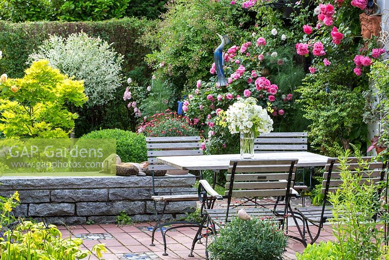 Wood and iron garden furniture on a brick stone terrace in a pottery maker's garden with ceramic objects and Rosa 'Leonardo da Vinci', 'New Dawn', 'Rosarium Uetersen', Acer japonicum 'Aconitifolium', Buxus and Sarracenia