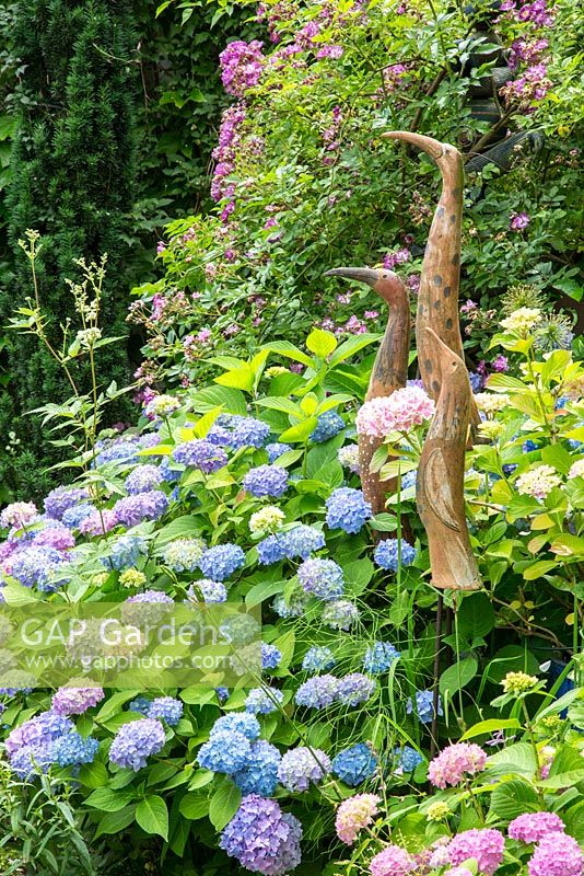 Three long shaped ceramic birds next to Rosa 'Veilchenblau' and Hydrangea macrophylla 'Endless Summer'