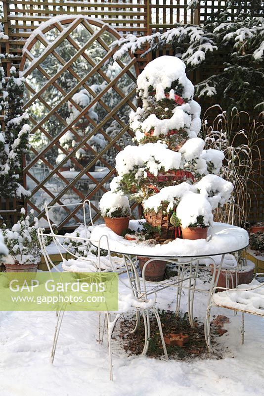 Courtyard with seating area and pots in winter. Picea glauca var. albertiana 'Conica' in pot on table with decorations made of red ribbon. A mirror trompe l'oeil creates a focal point on a wall of a garage - Welsch Garden, Berlin, Germany