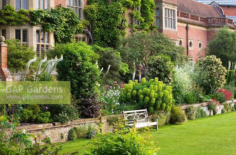 View to the opera house over the double herbaceous borders with white foxtail lilies - eremerus and euphorbia - wooden bench on lawn. Glyndebourne, East Sussex