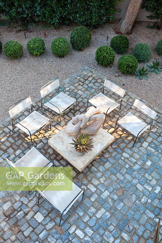 View of cobbled seating area and gravel garden with wooden sculpture in the sun and shadows. USA, August.