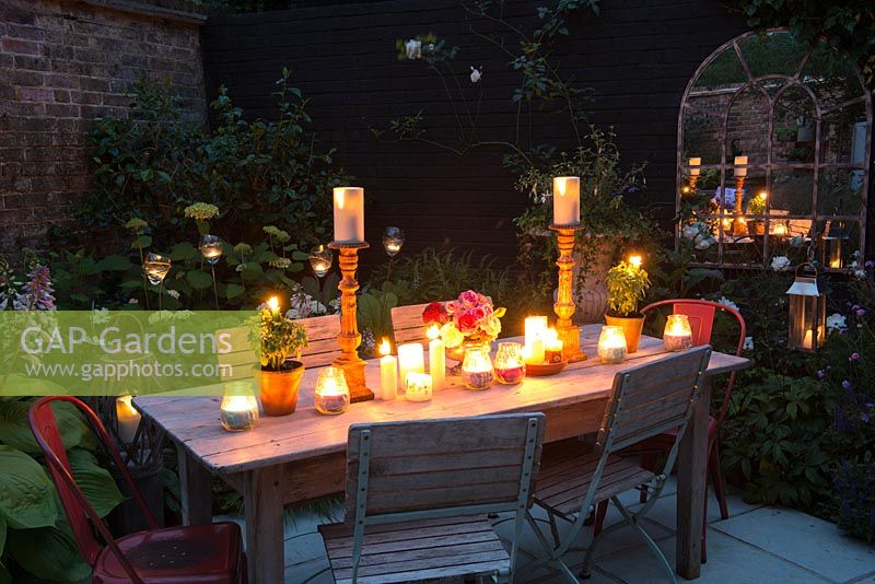 Table is prepared for summer barbecue with vase of roses and candles. Glass candleholders in borders illuminate planting from left to right, hosta, epimedium, Hydrangea 'Annabelle', lysimachia, Cosmos 'Purity', Verbena bonariensis. Reflections in wall mirror to far end of courtyard.
