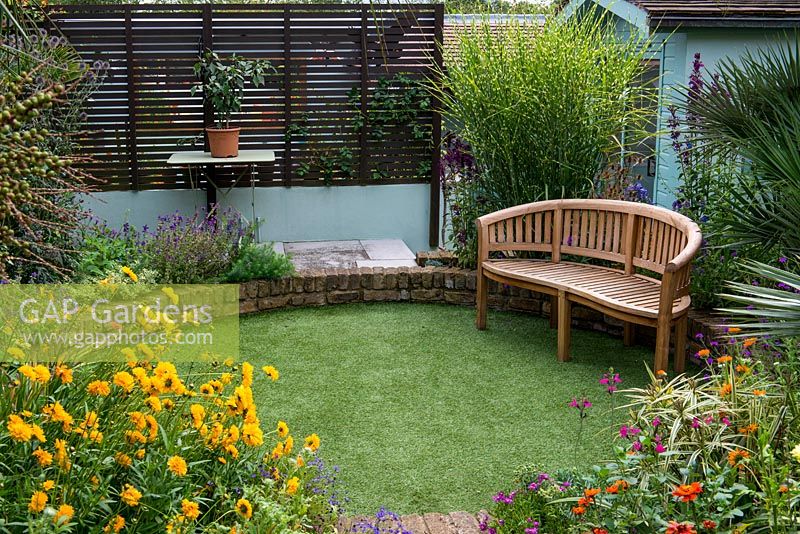 A sunken circular lawn of artificial grass, with a wooden bench in front of miscanthus sinensis 'Zebrinus'.