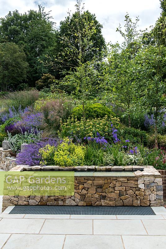 A raised stone water feature with bank planted with perennials, trees and ornamental grasses including: Campanula persicifolia, Alchemilla mollis, Phlomis russeliana, flag iris, Betula nigra and Molinia 'Karl Foerster'