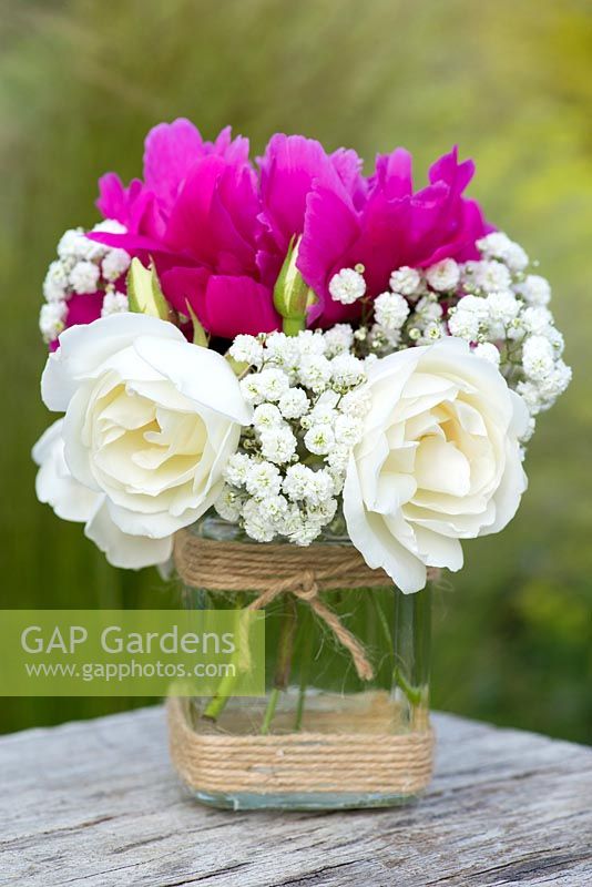 A summer posie with white roses, gypsophila and pink peony in a glass jar decorated with twine.