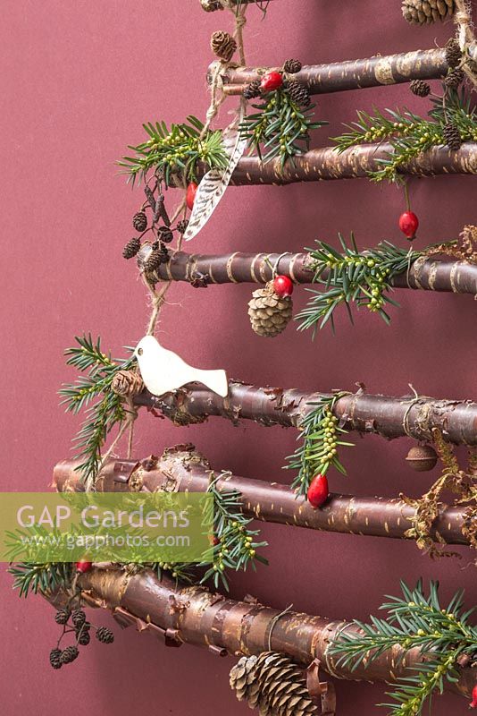 A hanging Christmas tree made with Birch branches, against a red background. Decorations feature Pine cones, feathers, Rose hips and Pine foliage