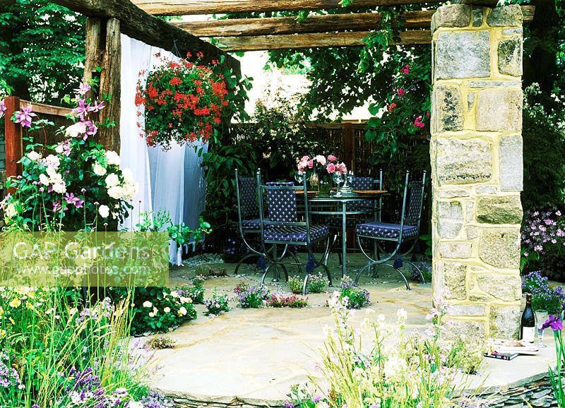 Patio with furniture, Hampton Court 2001, Design: Paul Dyer, the sound of the  countryside. Crazy paving patio with table and chairs, old timber pergola, geranium hanging basket, clematis, rosa and verbena  bonariensis