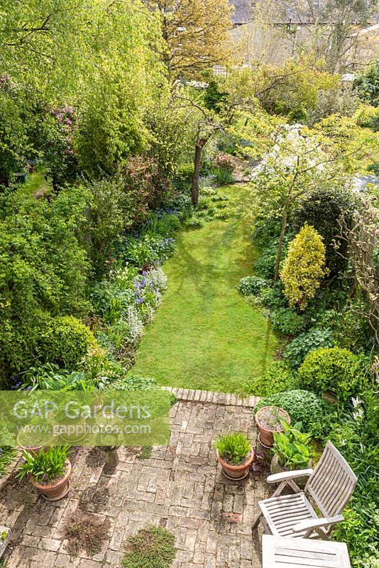 Aerial view of long, narrow, town garden in spring with informal lawn and mixed borders. Reclaimed brick patio with hostas and agapanthus in containers.