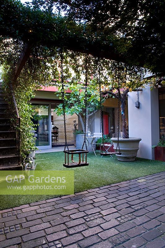 Courtyard garden with steps up to balcony, overhead steel-girder pergola with climbing plants and two swings, astroturf and plants in large containers. Modern house conversion of a disused factory.