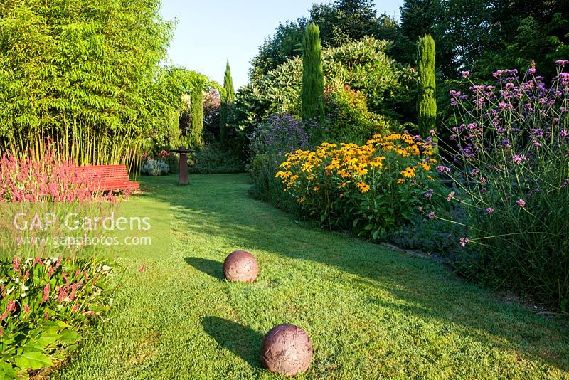 Bench on lawn backed by bamboo and borders featuring Verbena bonariensis, Rudbeckia, Euphorbia characias, Persicaria amplexicaulis and Cupressus sempervirens - July, Les Jardins de la Poterie Hillen