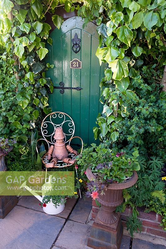 Green painted trompe l'oeil doorway in garden boundary with hedera colchica ivy and a white seat with a flowerpot man ornament and an old enamel watering can. 