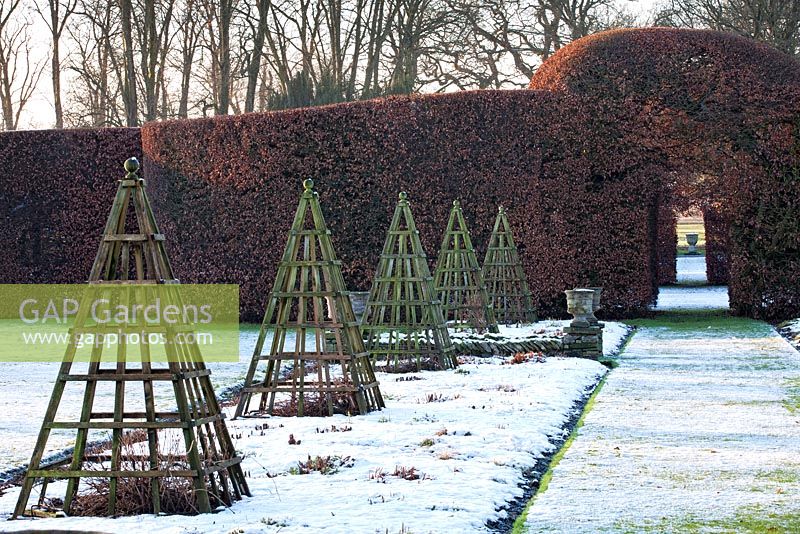 Herbaceous borders with wooden pyramid obelisks covered in snow at Levens Hall and Garden, Cumbria, UK. 