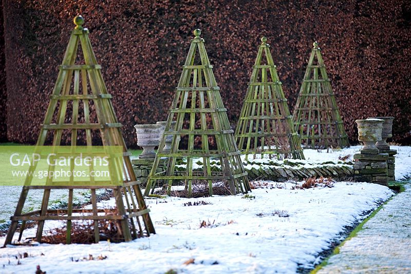 Herbaceous borders covered in snow with wooden pyramid obelisks at Levens Hall and Garden, Cumbria, UK. 