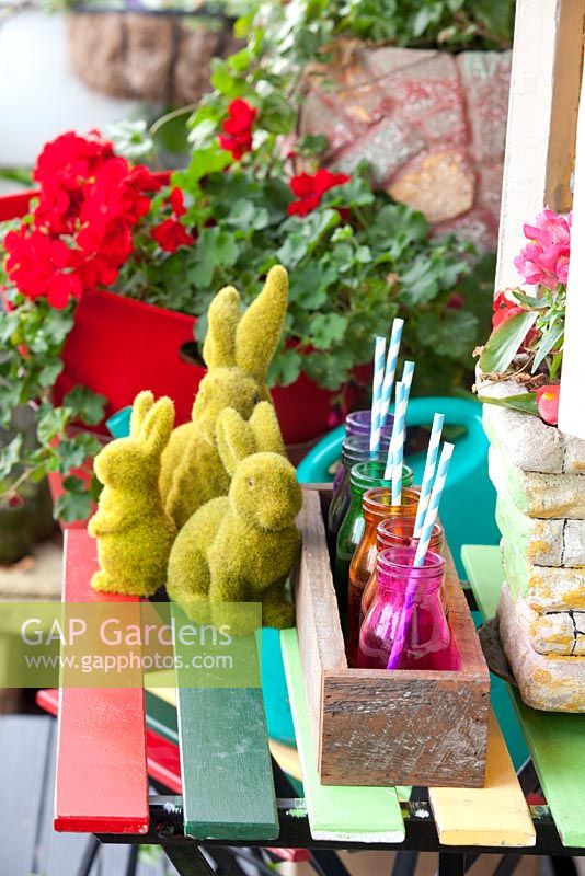 Detail of an inner city garden with three artificial grass rabbit ornaments, retro cement pots, recycled timber box with coloured glass bottles with drinking straws and colourful outdoor table and chairs.